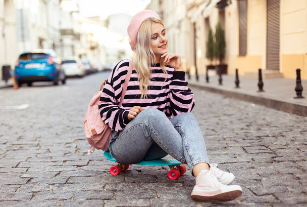 Молода класна дівчина в береті з рюкзаком сидить на скейтборді на європейській вулиці. - Фото, зображення