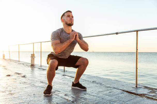 Athletic man practicing squats in the early morning on the beach - Valokuva, kuva