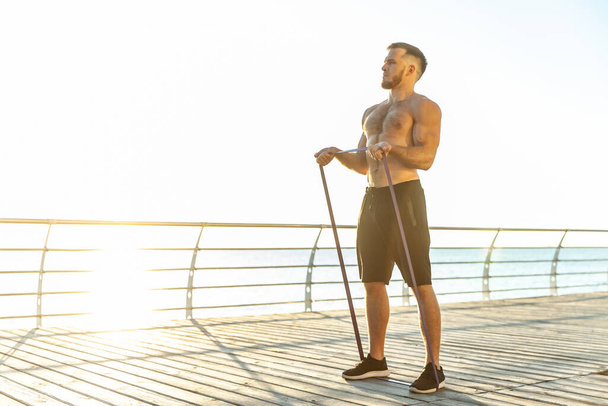 Νεαρός αθλητικός άνδρας με γυμνό κορμό γυμνάζεται με λαστιχάκια γυμναστικής νωρίς το πρωί στην παραλία. Μυϊκή εκπαίδευση. Υγιής τρόπος ζωής - Φωτογραφία, εικόνα