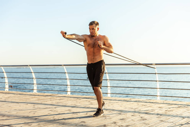 Young athletic man with a naked torso works out with fitness rubber bands in the early morning on the beach. Muscle training. Healthy lifestyle - Photo, Image