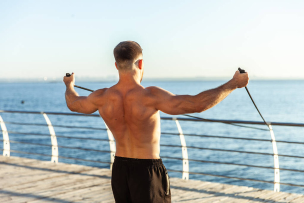 Fiatal atléta férfi meztelen felsőtesttel kondizik fitnesz gumiszalagokkal kora reggel a parton. Izomtréning. Egészséges életmód - Fotó, kép