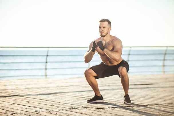 Atletický muž cvičí dřepy v časných ranních hodinách na pláži - Fotografie, Obrázek
