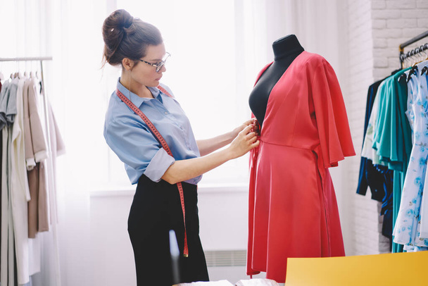 Side view of pensive woman seamstress with measuring tape standing near mannequin taking measurements while sewing new fashion dress in modern atelier - Foto, imagen
