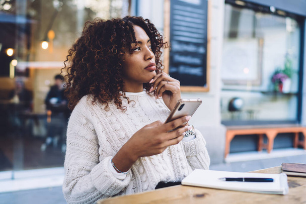 Mujer afroamericana pensativa con ropa casual sentada en la mesa de madera y mirando el teléfono móvil mientras descansa en la cafetería al aire libre - Foto, imagen