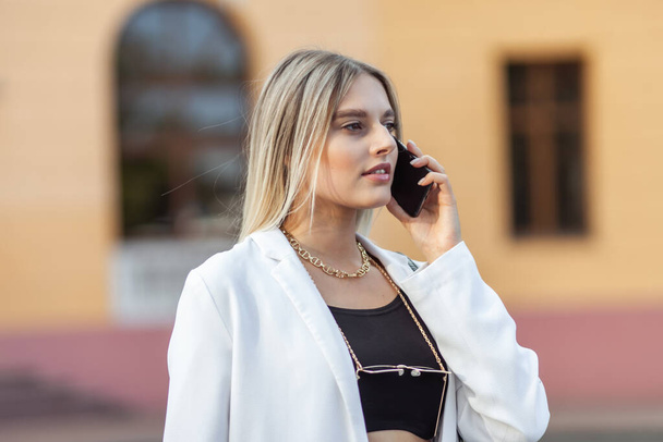 Young fashion woman in a white trendy jacket talking on phone in the city. Street fashion - Photo, image