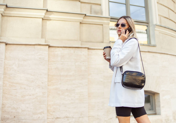 Молодая блондинка в модном наряде пьет кофе по дороге и разговаривает по телефону в городе - Фото, изображение