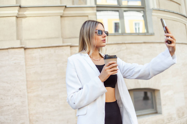 Joven mujer rubia de moda en traje de moda bebe café sobre la marcha y hace selfie en el teléfono en la ciudad - Foto, imagen
