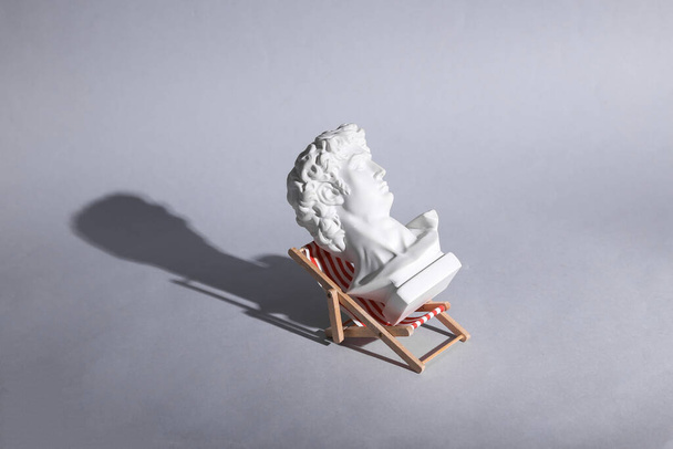 Perfect vacation concept. Bust of David rests on beach deck chair. Gray background. Minimal still life - Photo, image