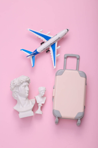 Bagaglio giocattolo con aereo aereo e busto David su sfondo rosa. Disposizione creativa. Minima natura morta da viaggio. Piatto. Vista dall'alto - Foto, immagini