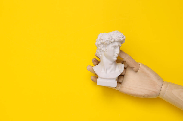Wooden hand holding David bust on yellow background. Minimal still life - Foto, Imagem
