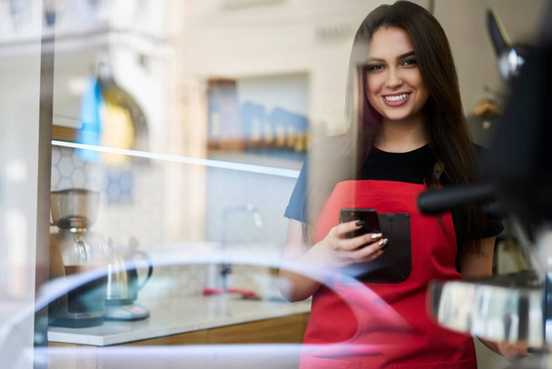 Half length portrait of cheerful caucasian female waitress in apron using mobile phone for making booking via app in cafeteria - Fotoğraf, Görsel