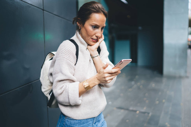 Side view of confused adult female in casual clothes and backpack smiling and browsing smartphone while standing near modern building - Foto, imagen