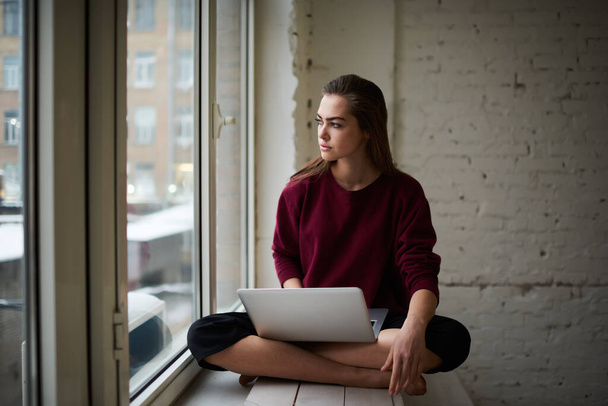 Serious female remote worker in casual clothes looking away sitting on windowsill with crossed legs and using opened laptop while working - Foto, imagen