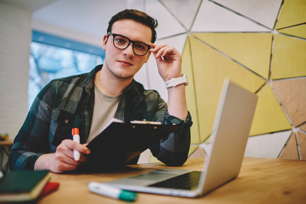 Concentrated handsome male in casual clothes looking at camera touching eyeglasses and sitting at table with opened laptop and clipboard while working - Фото, изображение