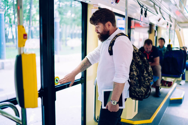 Side view of young man with beard in casual clothes standing in modern train and looking away in city in soft daylight - Valokuva, kuva