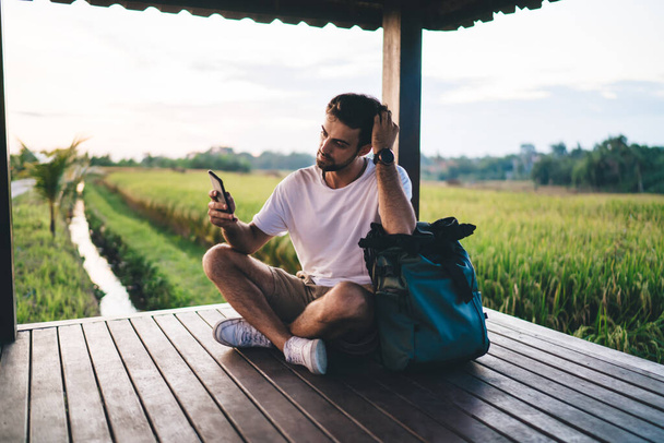 Full body emotionless bearded male wearing casual outfit sitting with legs crossed in wooden alcove and surfing modern mobile phone while trekking - Foto, Bild