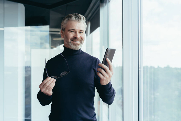 Portrait of a successful gray-haired man, senior businessman boss in the office smiling and looking at the camera by the window, holding a phone and glasses - 写真・画像
