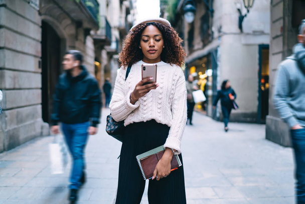 Serious dark skinned woman in trendy wear walking on street share content browsing web pages on smartphone - Fotografie, Obrázek