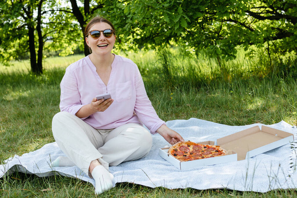 Happy young woman eating pizza on nature background and browsing internet on phone. Portrait of stylish beautiful woman using mobile phone reading text message outdoors. Fast food banner - Zdjęcie, obraz
