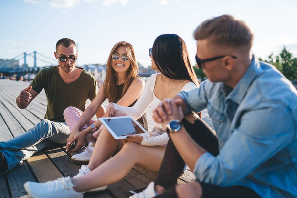 Group of young people in casual clothes and fashionable glasses talking while sitting in park with bridge and sky on background - Фото, зображення
