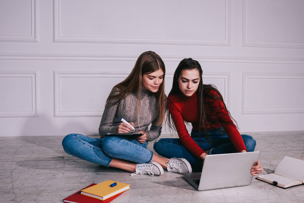 Young female students in casual outfit sitting on floor with legs crossed and browsing laptop while doing homework in light room - Zdjęcie, obraz