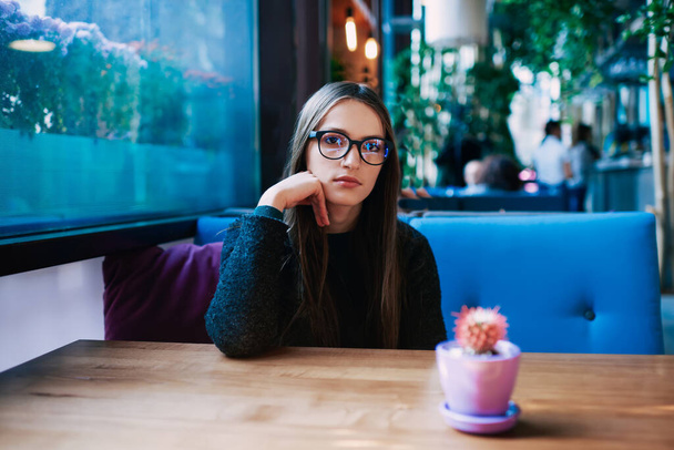 Wistfl female in casual clothes with eyeglasses looking at camera while sitting at wooden table with cactus in flower pot leaning  - Photo, image