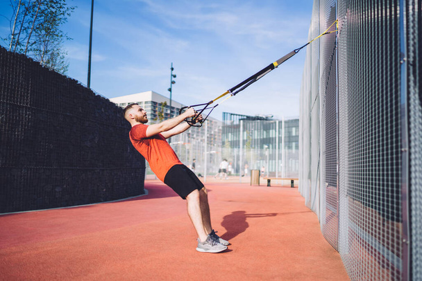 Side view of full body powerful concentrated male athlete wearing sportswear doing suspension training on sports ground on sunny day - Fotografie, Obrázek