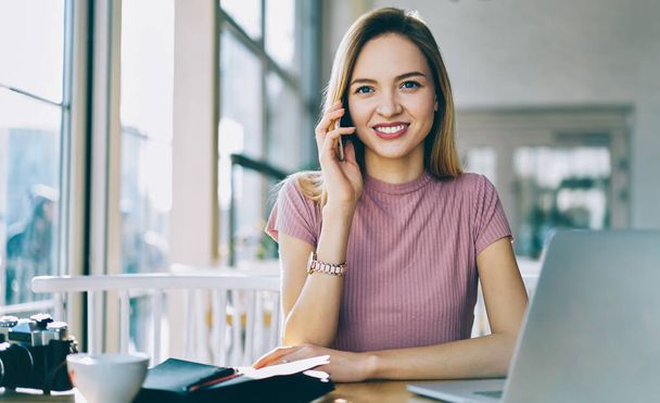 Portrait of cheerful woman using application for talking with friend via smartphone working remotely at laptop device - Fotoğraf, Görsel