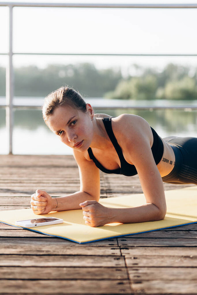 Retrato de estilo de vida de verano de una hermosa joven rubia feliz con un cuerpo sexy bronceado en ropa deportiva haciendo ejercicios en una esterilla de yoga - Foto, imagen