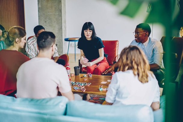Multiracial group of young friends in casual clothes sitting at wooden table loaded with playing cards tokens in cozy room - Photo, image