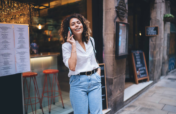 Half length portrait of cheerful female tourist smiling at camera while connecting to roaming internet for make international mobile talking in city - 写真・画像