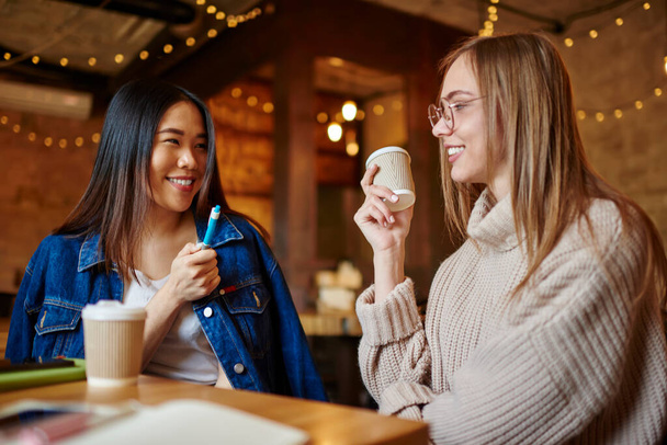 Fröhliche ethnische Frau mit zahmem Lächeln sitzt am Tisch und kommuniziert mit positiver Freundin mit Kaffee zum Mitnehmen Zeit im Café verbringen - Foto, Bild