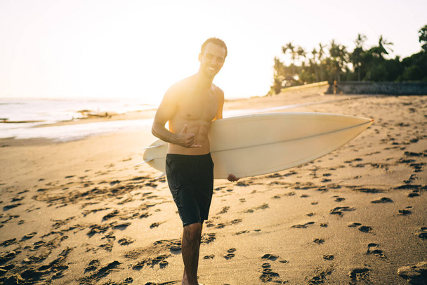 Happy young shirtless male surfer in swimwear with shortboard smiling and doing shaka sign with hand while recreating on sandy beach against bright sunlight - Photo, image