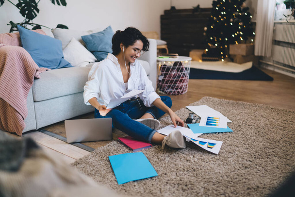 Full length smiling young female in jeans and white shirt sitting on floor with papers and laptop while listening to good music via earphones - Zdjęcie, obraz