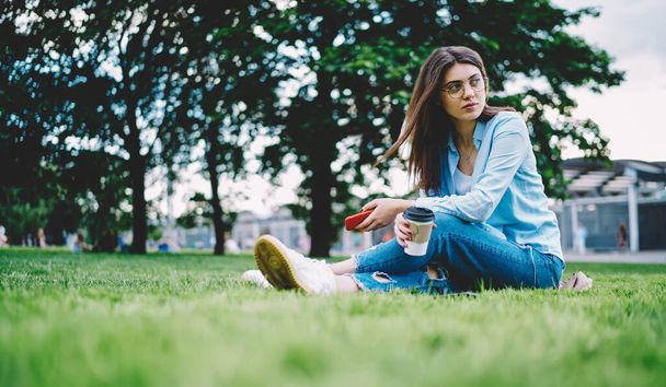 Chica hipster contemplativa con taza de café para llevar y dispositivo de teléfono inteligente en manos reflexivas mirando hacia otro lado mientras descansa en el parque - Foto, imagen