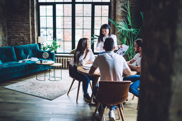 Positive young multiracial coworkers wearing blue jeans and white shirts gathering at table and sharing ideas while working together in creative coworking space - Foto, imagen