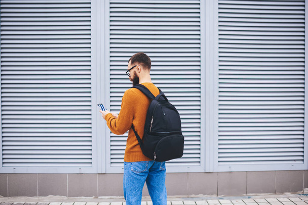 Back view of slender young bearded hipster with black backpack checking notifications on mobile phone beside modern building on street - 写真・画像