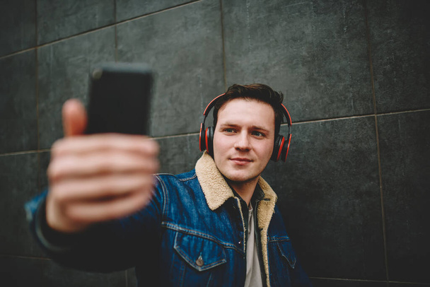 Glad handsome male with headphones listening to music taking selfie on camera of mobile phone while standing near wall on street - Photo, Image