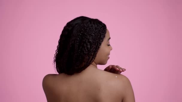 Cuidado corporal y nutrición. Vista trasera de la joven bastante irreconocible dama afroamericana aplicando crema de mimos en su hombro desnudo, posando sobre fondo de estudio rosa, cámara lenta - Metraje, vídeo
