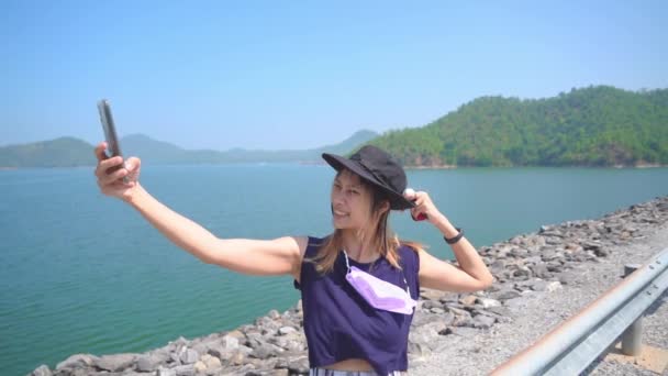 Asiática turista mujer está tomando foto selfie en la orilla de un frente de agua con lago e isla en el fondo. - Imágenes, Vídeo