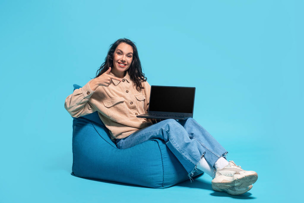 Cheerful funny young caucasian lady brunette digital nomad, student sitting on bagchair and pointing finger at laptop with empty screen isolated on blue background. Vlog, social media, ad and offer - 写真・画像