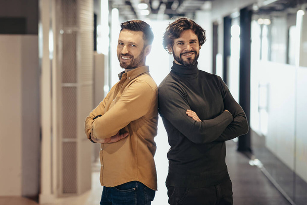 Portrait of happy handsome entrepreneurs posing with crossed arms and smiling in company hall. Confident cheerful businessmen standing back to back at contemporary office. Business partnership concept - Φωτογραφία, εικόνα
