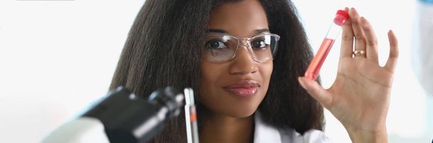 Une femme à la peau foncée dans un laboratoire tient un échantillon avec un liquide rouge. Test sanguin, analyse d'ADN, recherche de virus - Photo, image