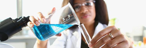 Científica de piel oscura vertiendo líquido azul en un tubo de ensayo, de cerca, borroso. Experimentar con drogas - Foto, Imagen