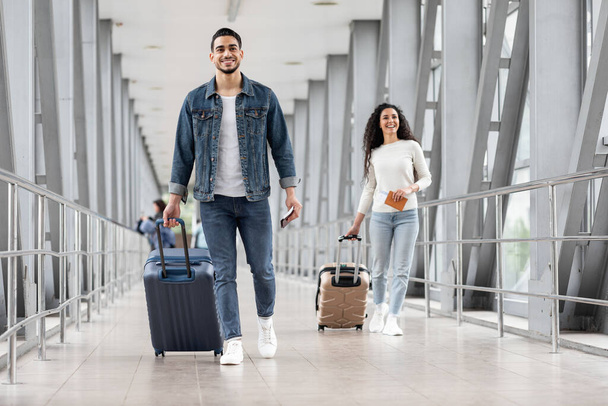 Ulaşım konsepti. Havaalanında bavulları olan gülümseyen Arap adam ve kadın, mutlu erkek ve kadın valizleriyle çıkış kapısına gidiyor, hava yolculuklarının keyfini çıkarıyor, - Fotoğraf, Görsel