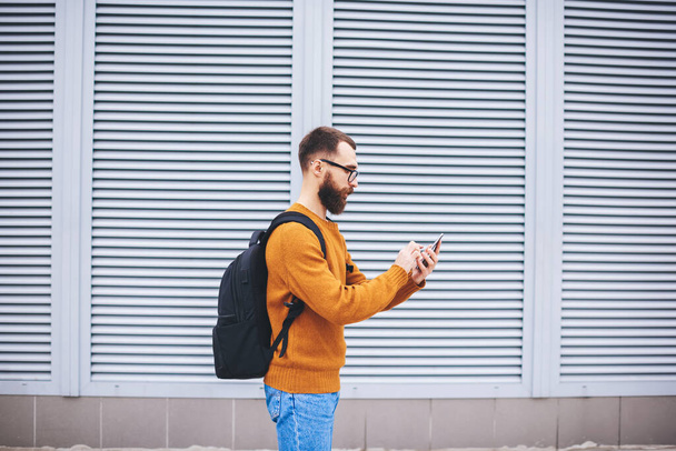 Вид сбоку на молодого бородатого хипстера в очках и рюкзачном наборе номера на мобильном телефоне рядом с домом с занавесками на окнах - Фото, изображение