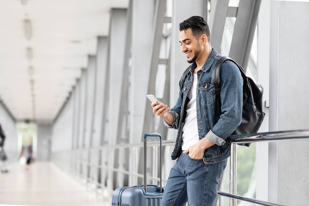 Portrét usměvavý arabský muž cestovatel s Smartphone a zavazadla čeká na letišti, šťastný mladý muž středního věku procházení internetu na mobilním telefonu, zatímco stojí v terminálu, kopírovat prostor - Fotografie, Obrázek