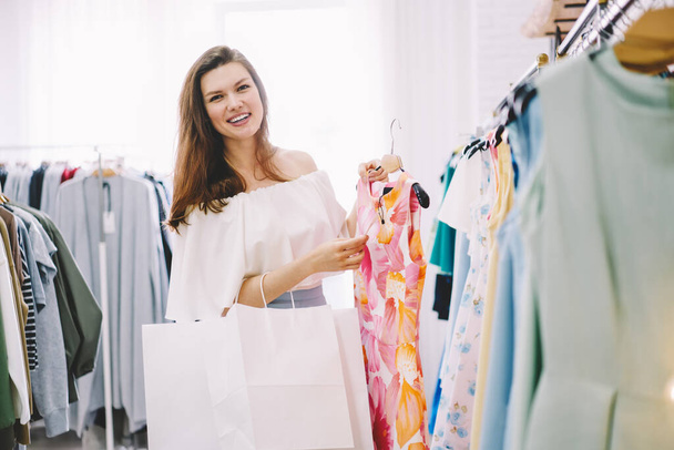 Cheerful brunette in white outfit with white shopping bags looking at camera and smiling while choosing dress for shopping in fashion boutique - Photo, Image