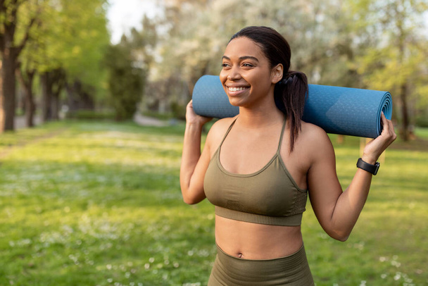 Portret van een vrolijke zwarte vrouw met een sportmat op haar schouders in het stadspark, vrije ruimte. Fit Afro-Amerikaanse dame klaar voor yoga praktijk, houden gehydrateerd tijdens outdoor workout - Foto, afbeelding