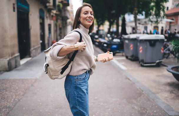 Cheerful female tourist with travel backpack standing at urban street and smiling satisfied with solo getaway vacations - Foto, Imagen